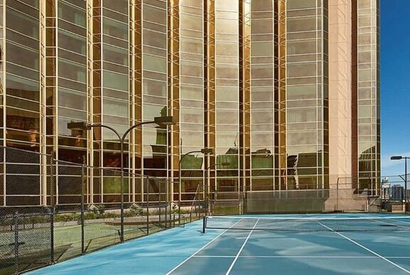 Tennis court overlooked by mirror-glass skyscraper with lighting.