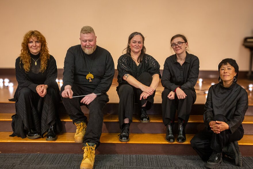Five musicians dressed in black, casually seated on low steps in a performance space.
