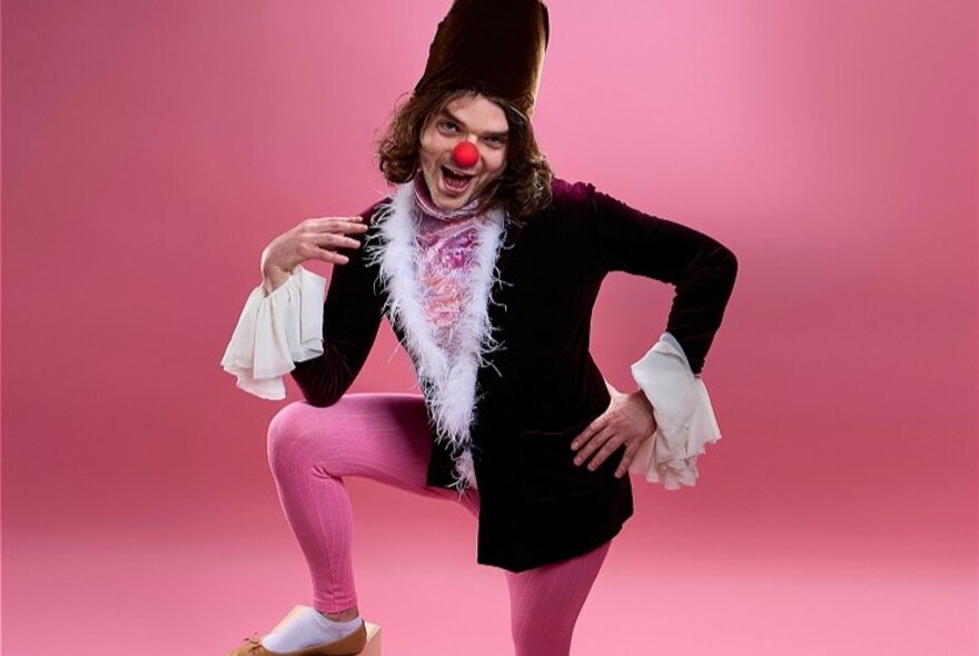 A man dressed up in a pink tights and an oversized black jacket with white frilly cuffs and lapels, a red nose and a tall hat.