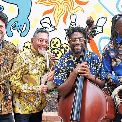 Four smiling musicians from Afrocubismo, wearing brightly patterned shirts and holding their instruments, posed against a wall mural.