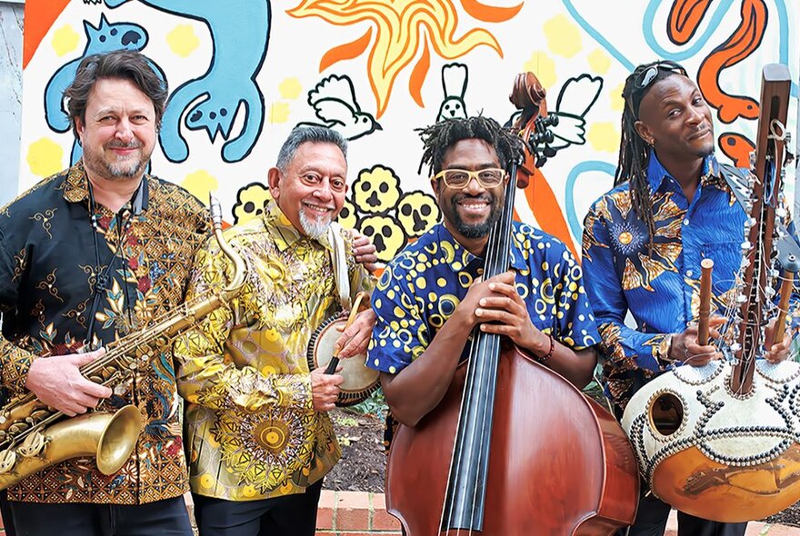 Four smiling musicians from Afrocubismo, wearing brightly patterned shirts and holding their instruments, posed against a wall mural.