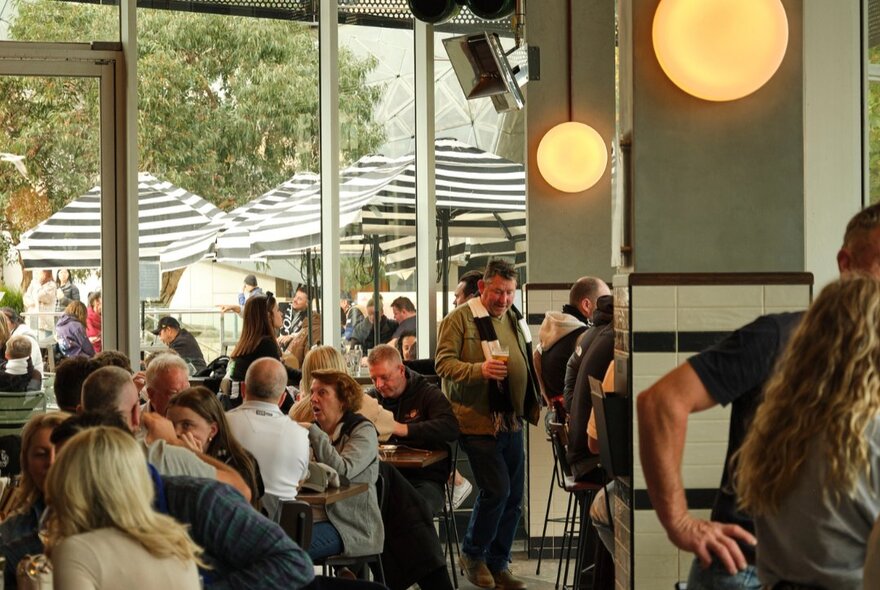 Interior of a pub with people socialising, featuring a large glass window wall that looks out onto an outdoor area with black-and-white striped umbrellas.