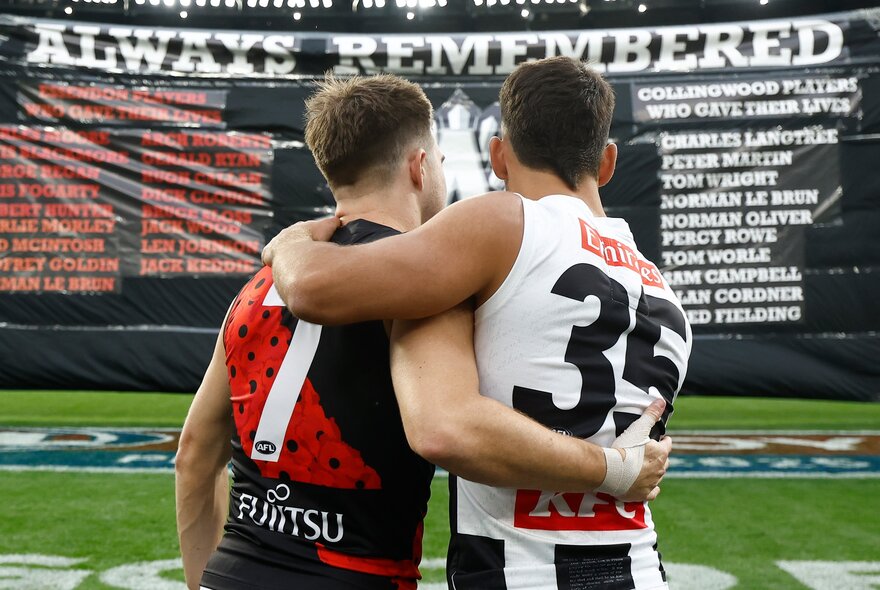 An Essendon and a Collingwood AFL player stand arm-in-arm in front of the ANZAC Day banner on the football field.