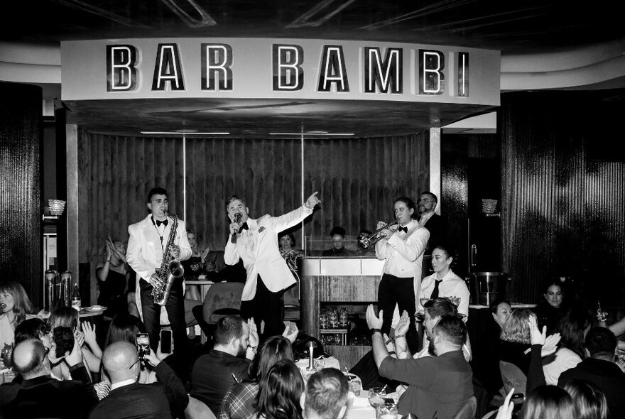 Jazz combo wearing white jackets performing under Bar Bambi signage in front of diners seated at tables.