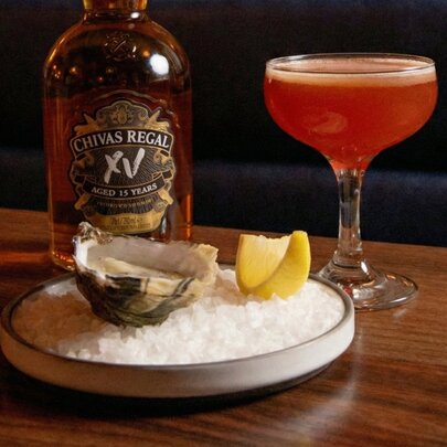 An orange-hued whisky cocktail, an oyster and slice of lemon on a plate and a bottle of whisky on a wooden table. 