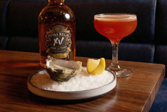 An orange-hued whisky cocktail, an oyster and slice of lemon on a plate and a bottle of whisky on a wooden table. 