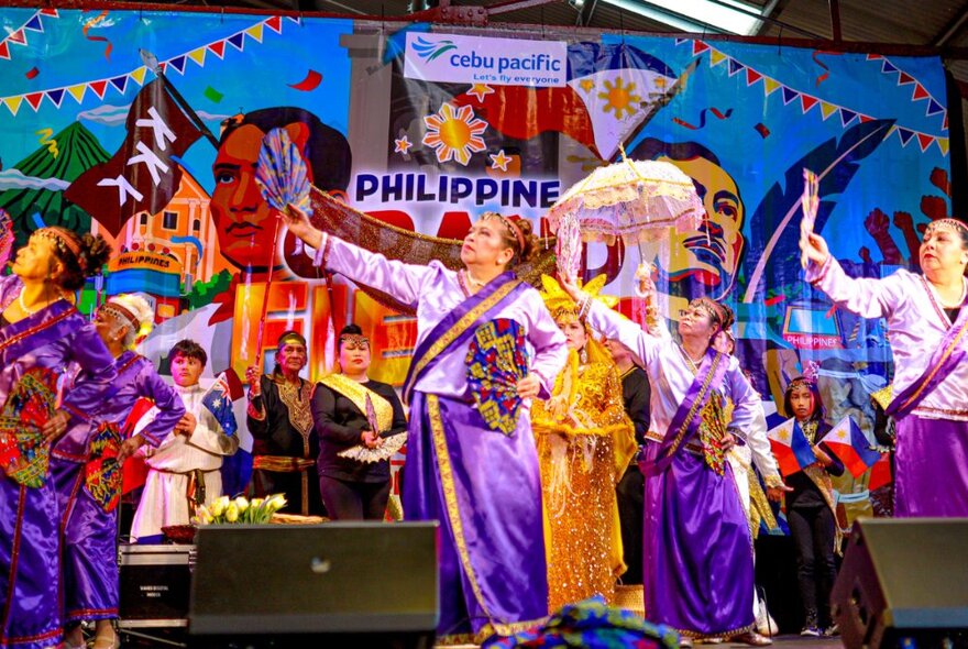 A colourful Filipino dance troupe on stage wearing traditional purple and gold outfits, in front of a bright painted backdrop.