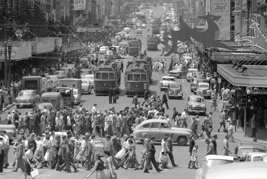 Black and white photograph of a busy Melbourne city intersection with trams, cars, people crossing the road, and buildings, dating from around the 1950s or 1960s.