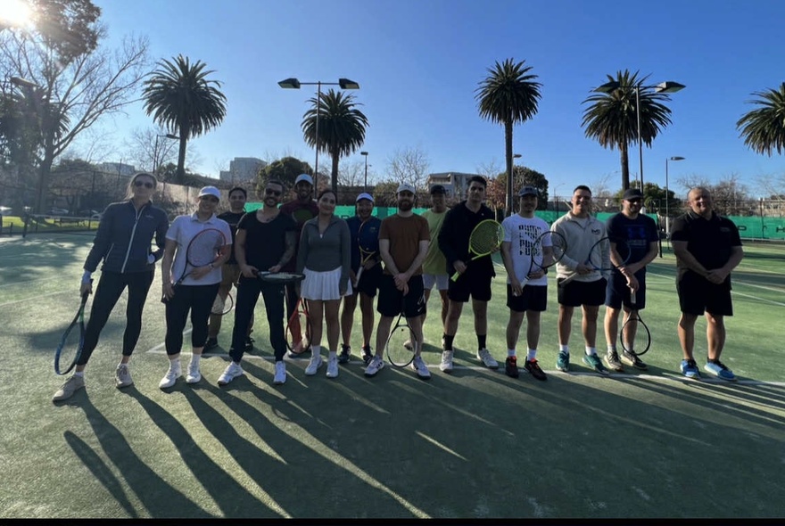 A group shot of people holding tennis racquets with palm trees behind them and long shadows in front of them. 
