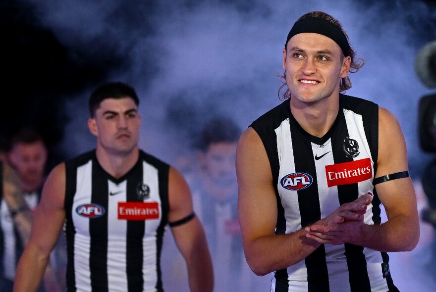 Two Collingwood football players with smoke from a smoke machine behind them.