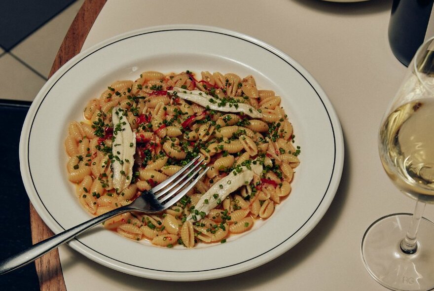 A fork resting in a dish of pasta next to a glass of white wine.