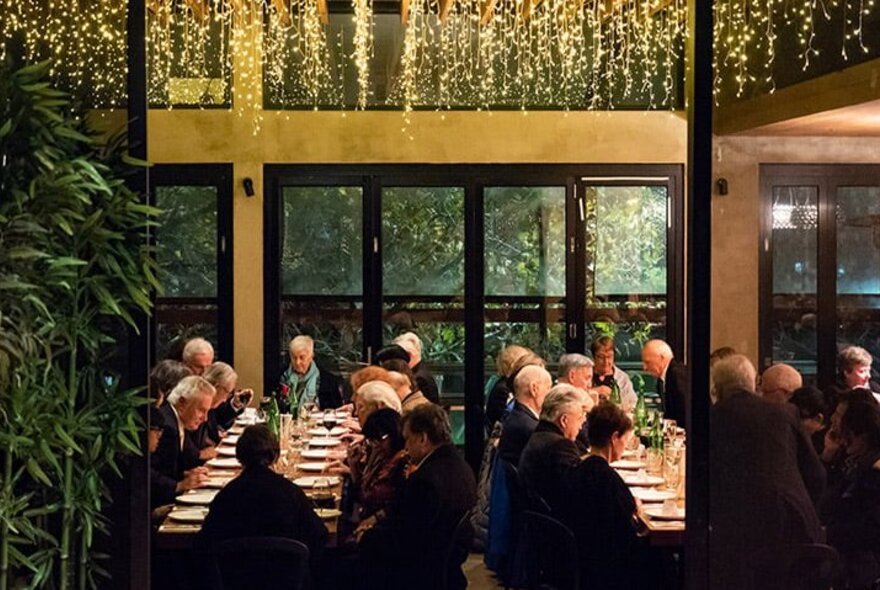 Diners sitting on long tables in a restaurant with gold fairty lights cascading down. 