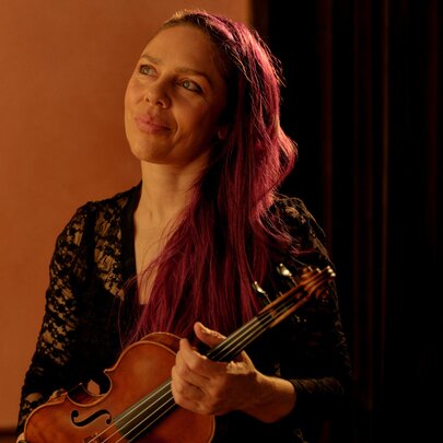 A woman with long red hair draped over one shoulder smiles as she holds a violin. 