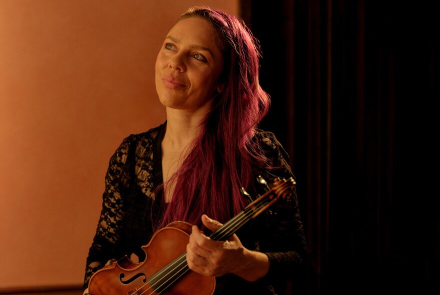 A woman with long red hair draped over one shoulder smiles as she holds a violin. 