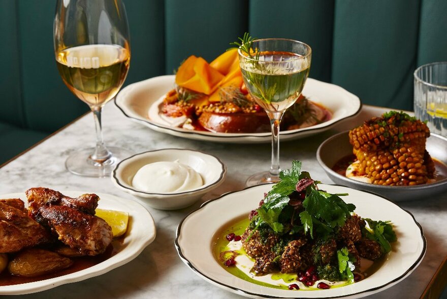 A table in a restaurant with plates of food, glasses of wine, and a banquette seat behind it.