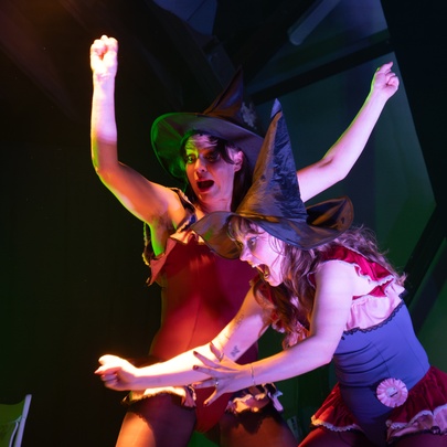 Two comedic performers in witches hats clowning around on a stage. 
