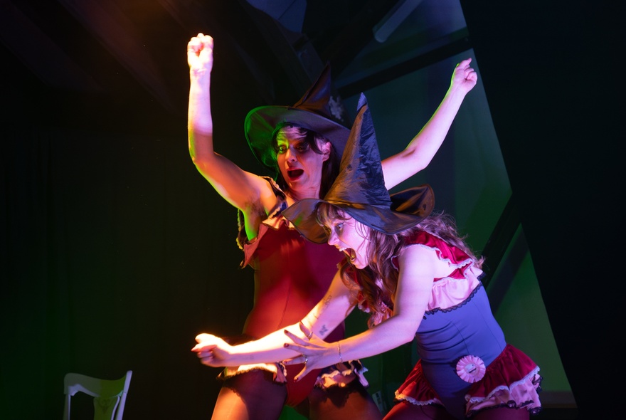 Two comedic performers in witches hats clowning around on a stage. 