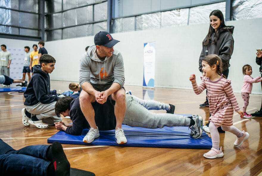 A person sitting on the back of a person holding a plank on an exercise mat in an indoor hall, surrounded by standing spectators.