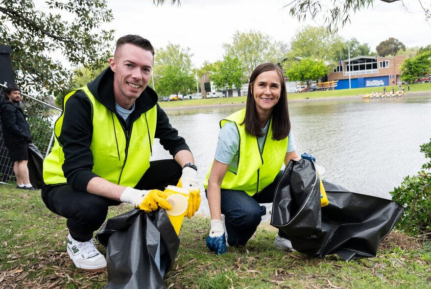 Two people wearing yellow high-visibility vests and gloves, picking up litter from the banks of the Yarra River and placing it into black garbage bags they are holding. 