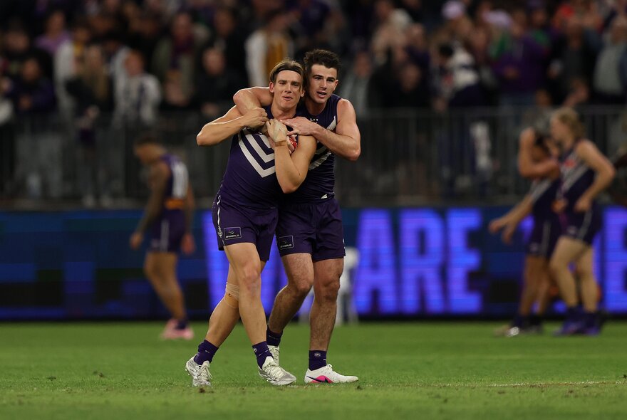 Two Fremantle Dockers AFL players celebrating on the field, one leaning into the other and hugging him from behind, a blurred stadium crowd behind them.