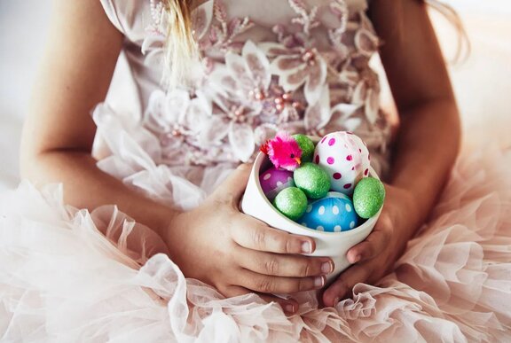 A child's hands holding a small white container of brightly coloured easter eggs in their lap.