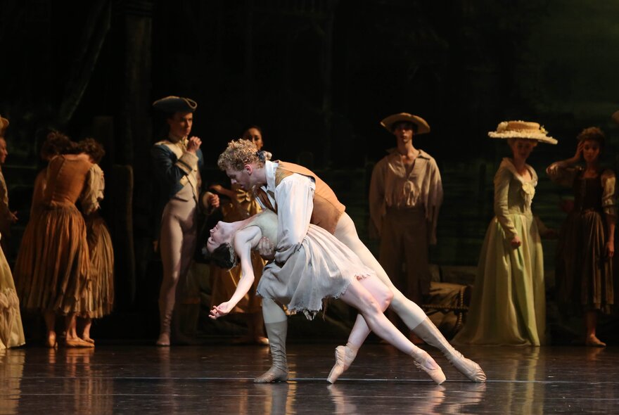 Ballet dancers performing on stage, dressed in 18th century style clothes, a central couple in an embrace with other dancers in the background shadows.