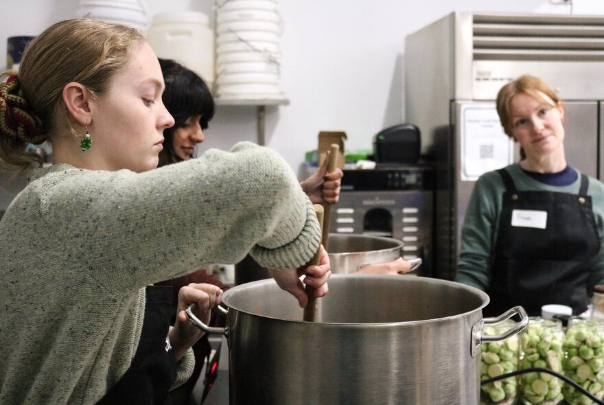 Cooking workshop with a person stirring a large pot in a kitchen setting with vegetables in pickling jars.