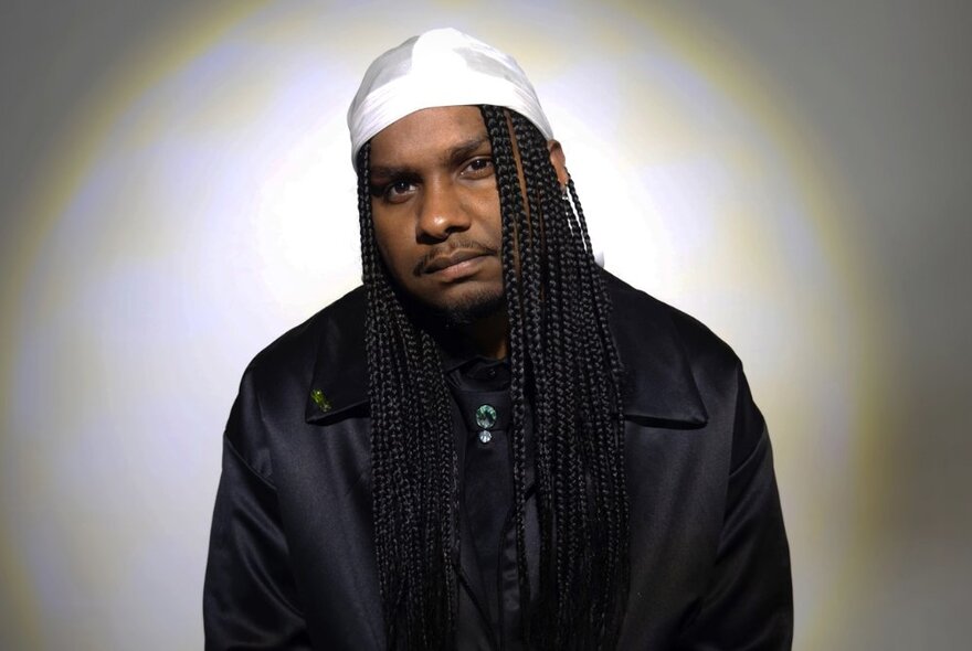 Singer and rapper Baker Boy, wearing a white cap over his long dark braided hair, and looking directly at the viewer.