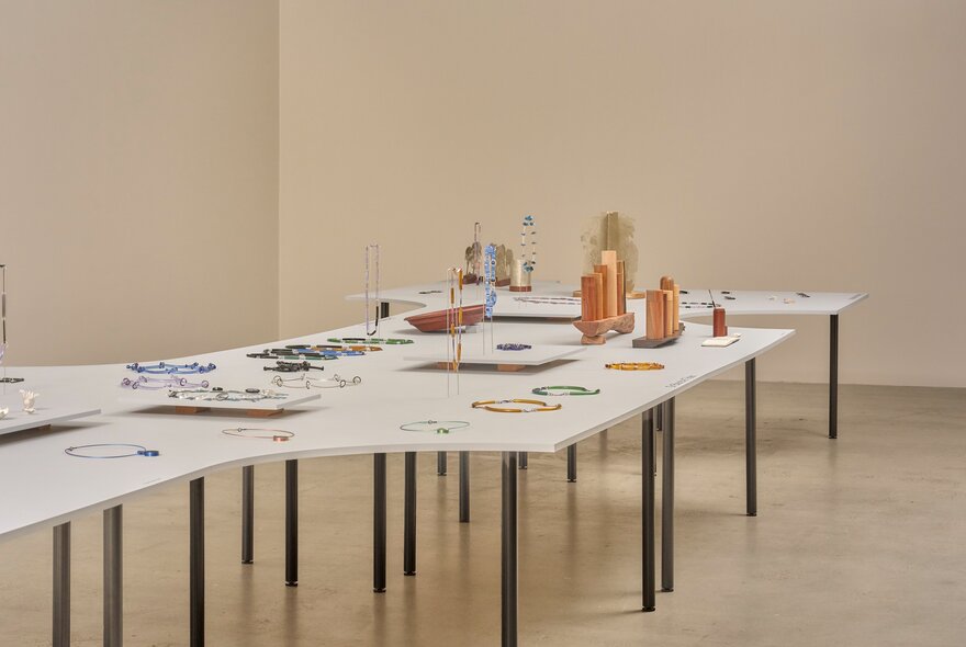 A gallery display featuring a series of long, white tables with slender black metal legs holding various decorative objects, jewellery and sculptural art objects.