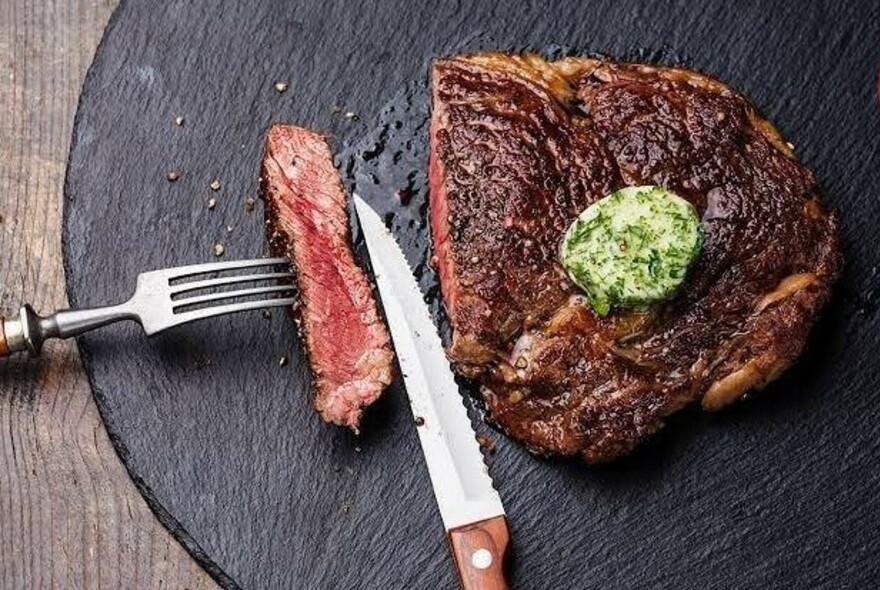 Knife and fork with slice of meat and large piece of steak on a black plate.