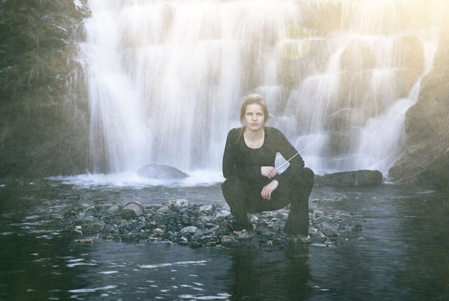 Orchestra conductor Tabita Berglund, wearing black and posed crouching outdoors in front of a waterfall.