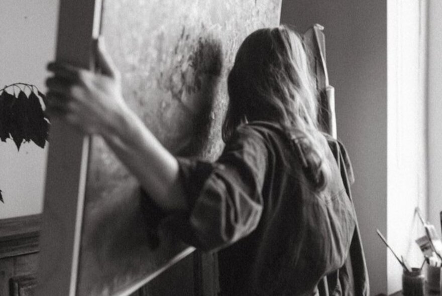 A black and white photograph of a woman moving a large framed canvas artwork.