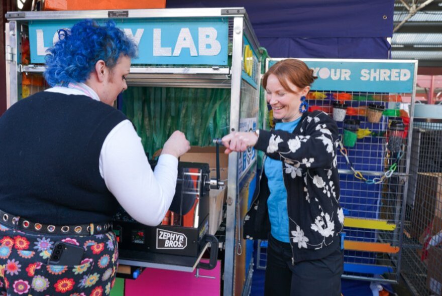 A person with blue hair at a vending machine as another person directs them on how to use it. 