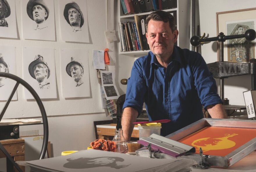 A portrait of artist, Clayton Tremlett, leaning on a workbench in his art studio with artworks of Australian First World War Digger Memorials hung on the wall behind him