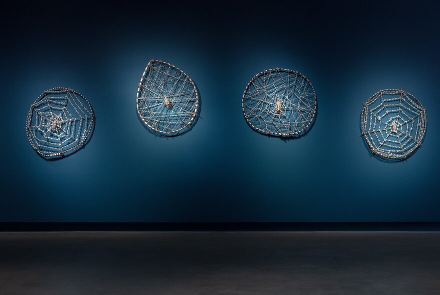 Installation view of an exhibition, with four flat sculptures hanging on a blue wall.