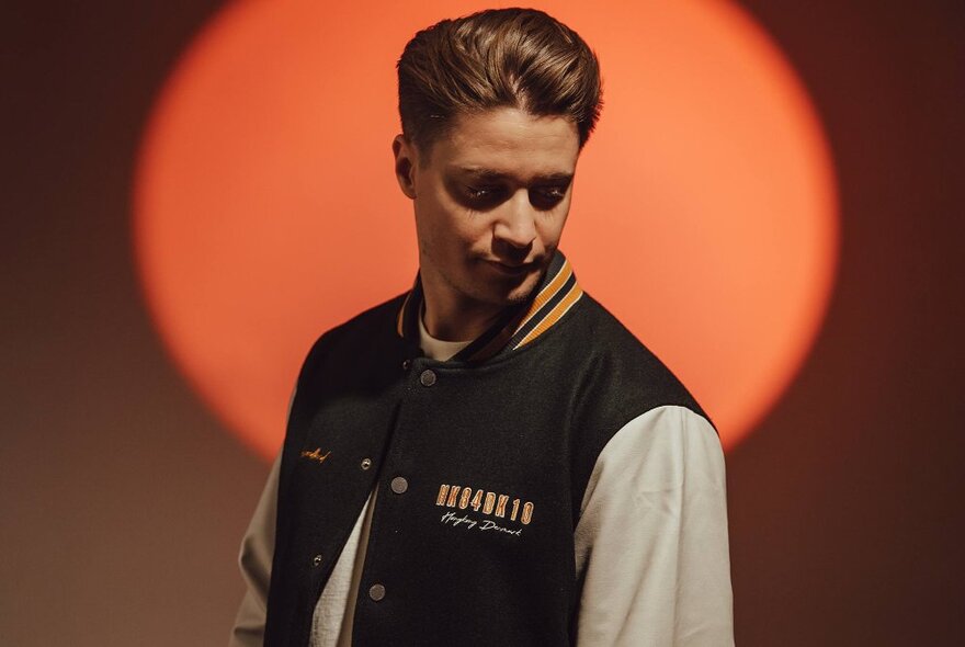 DJ Kygo in front of an orange light, wearing a logo baseball jacket.