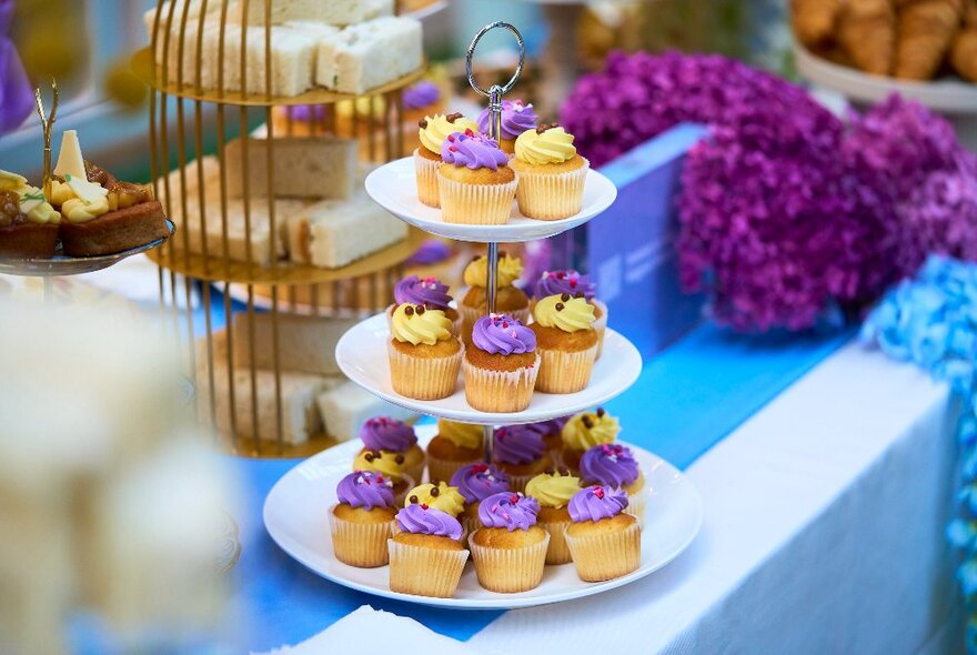 A three-tiered display plate of mini cupcakes, on a table decorated with purple and white flowers.