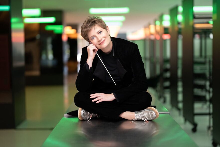 Conductor Anja Bihlmaier wearing a black suit and sitting cross legged on a steel tabletop, a baton in her hand, her head tilted to one side.