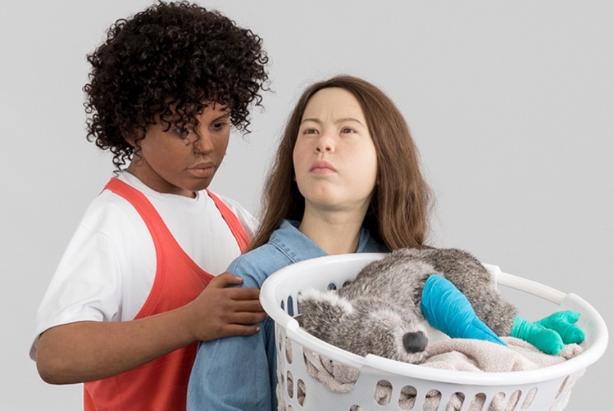 Close up detail of two figurative sculptures in a gallery space, carrying a plastic washing basket of laundry, in which a koala with bandaged feet and paws is resting.