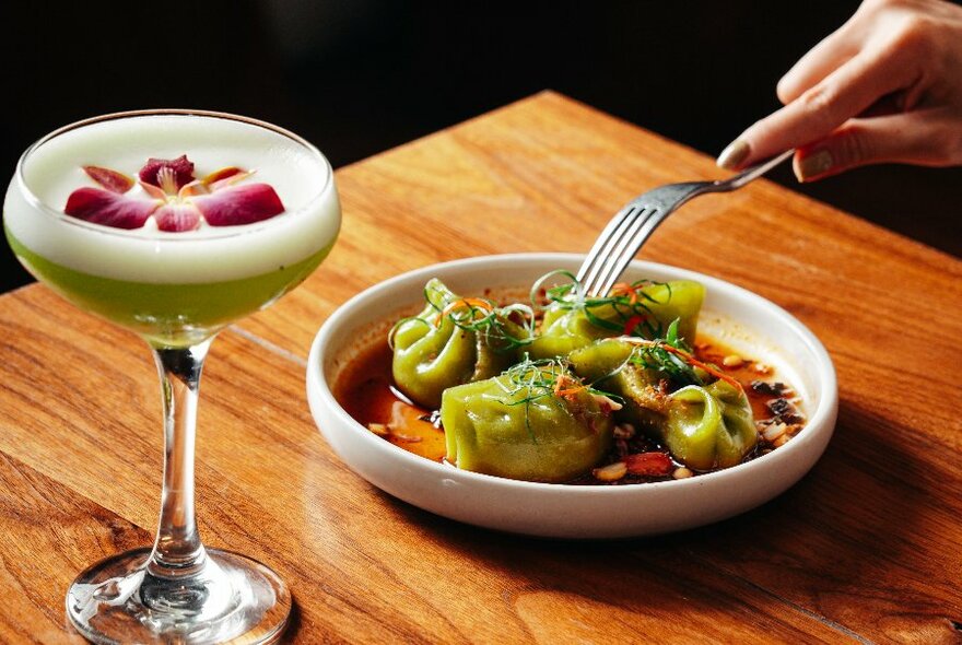 Cocktail next to plate of green dumplings in sauce with fork hovering over.