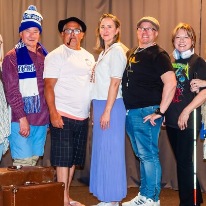 A local community theatre cast or group posing on stage with vintage suitcases, some dressed in costumes. 
