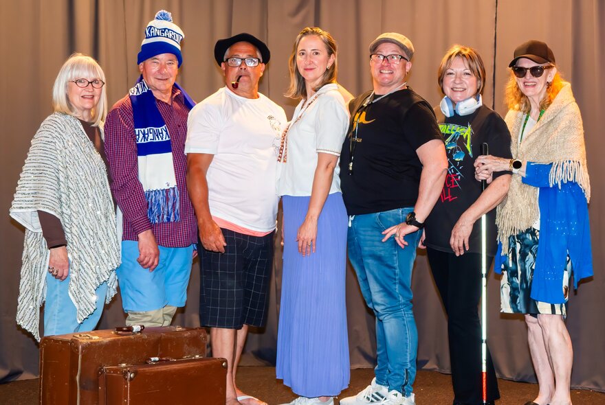 A local community theatre cast or group posing on stage with vintage suitcases, some dressed in costumes. 