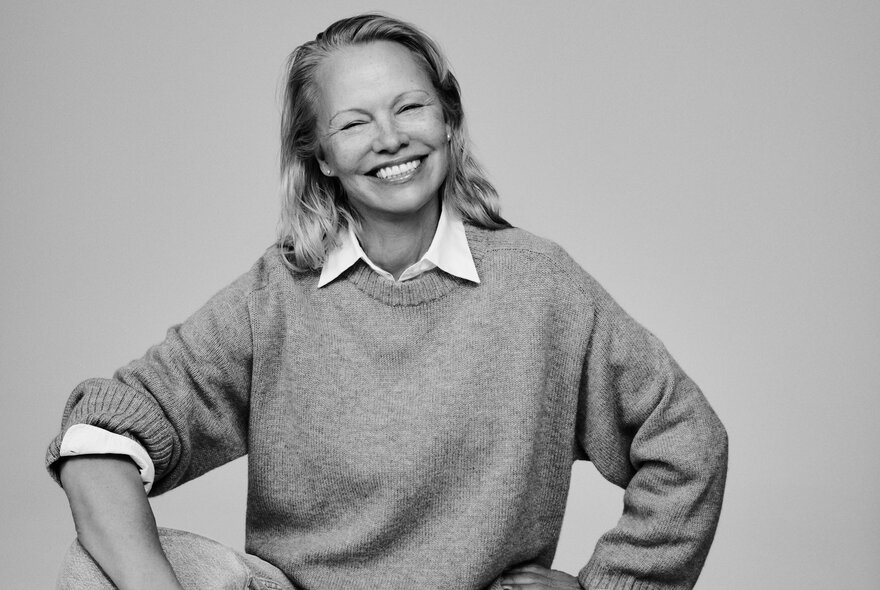 Black and white portrait of smiling 90s icon, Pamela Anderson, wearing a knit jumper over a white shirt.