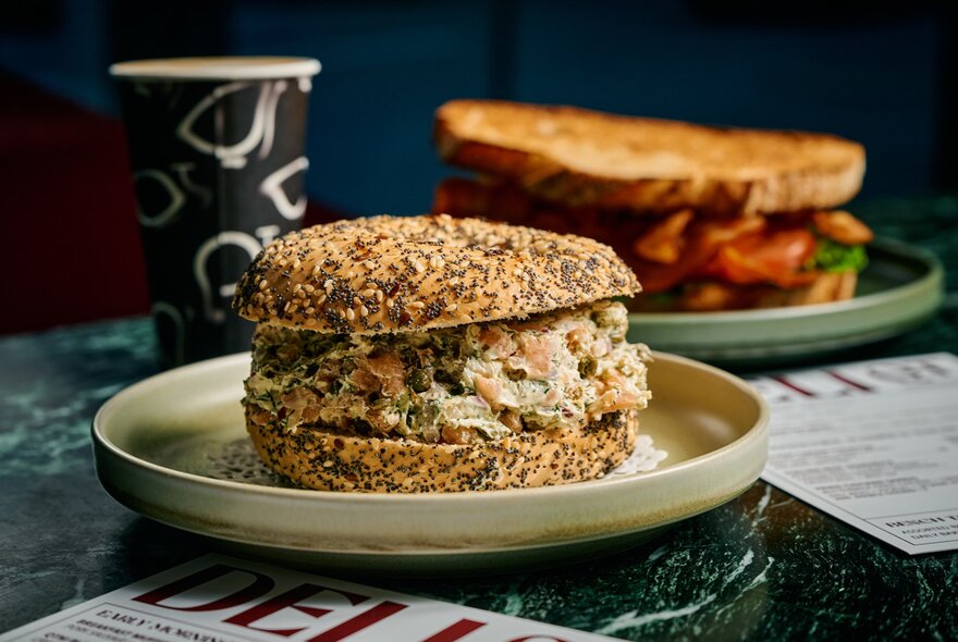 Snack-sized seed-crusted bagel sandwich on a plate, with a toasted sandwich and takeaway drink in the background.