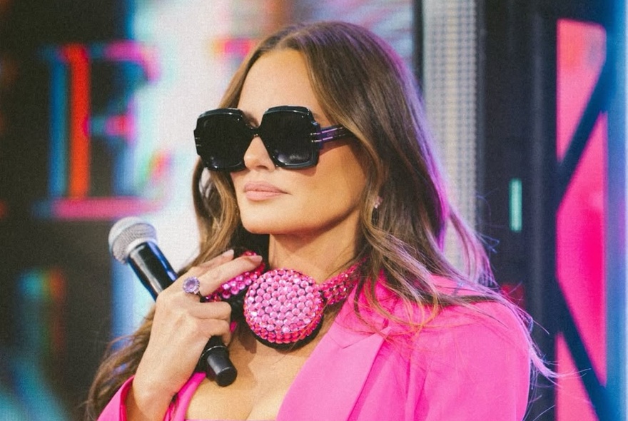 DJ Meredith Marks wearing sunglasses and dressed in hot pink and holding a microphone. 