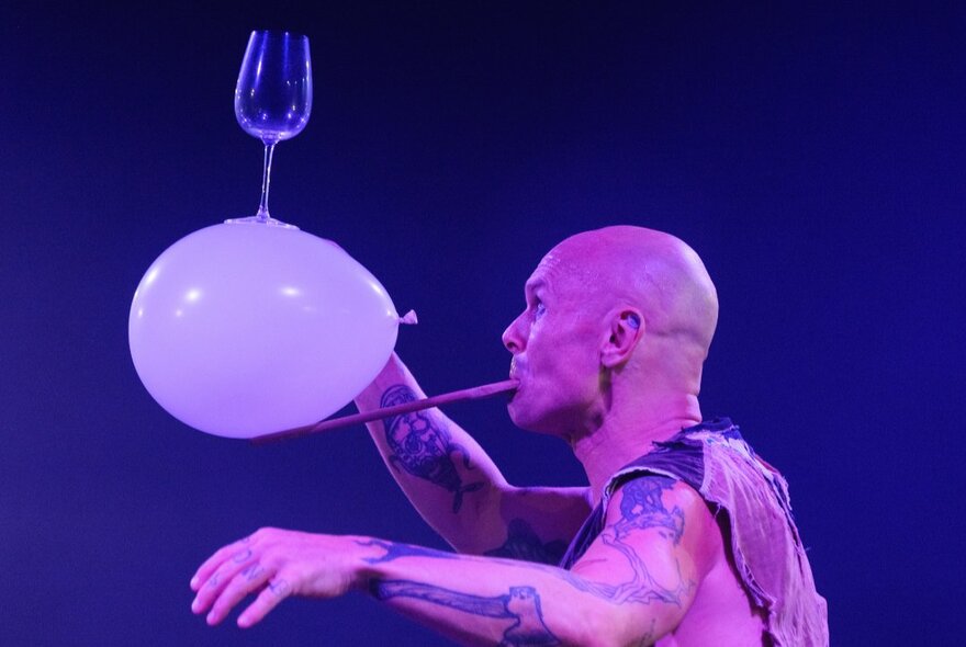 A man balanding a wine glass on a balloon, on a stick held in his mouth. 