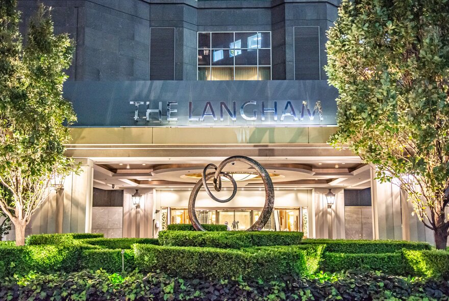 The illuminated entrance of The Langham hotel at night, featuring a metal sculpture and landscaped hedges in the foreground.