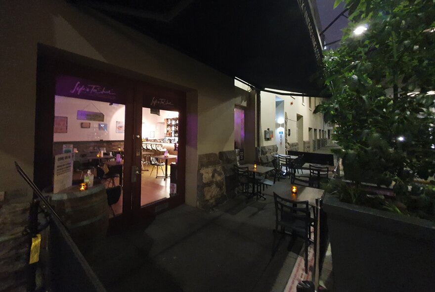 The courtyard at Life's Too Short bar at night, with tables and chairs, and a view through the glass doors into the interior space.