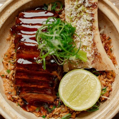 A clay pot filled with rice, topped with sliced glazed pork belly, bone marrow, a lime wheel, and fresh green garnish.