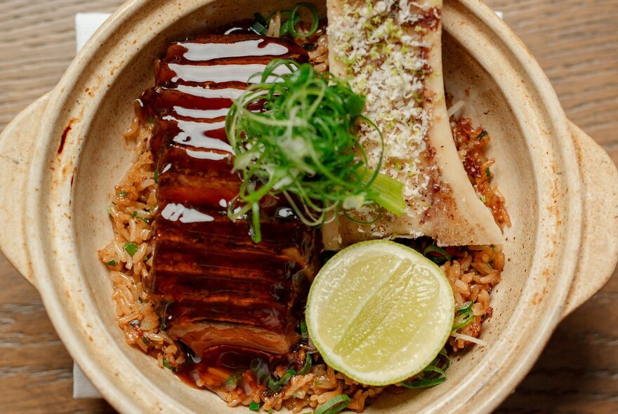 A clay pot filled with rice, topped with sliced glazed pork belly, bone marrow, a lime wheel, and fresh green garnish.